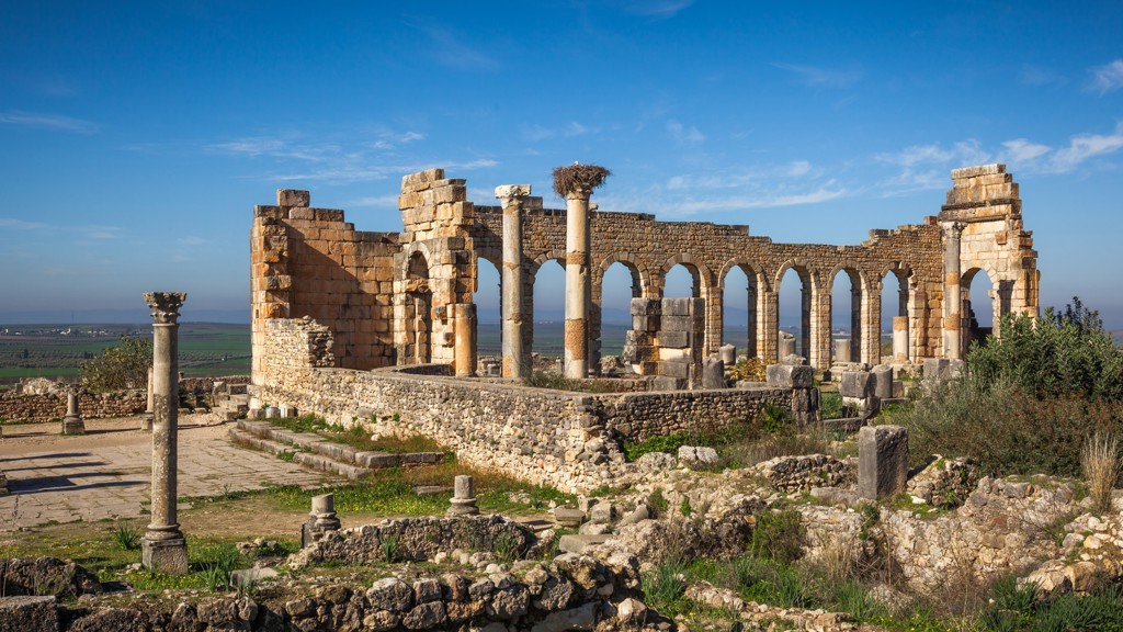Ruïnes van de Romeinse stad Volubilis langs de Route Marokko, met bogen en zuilen onder een blauwe lucht.
