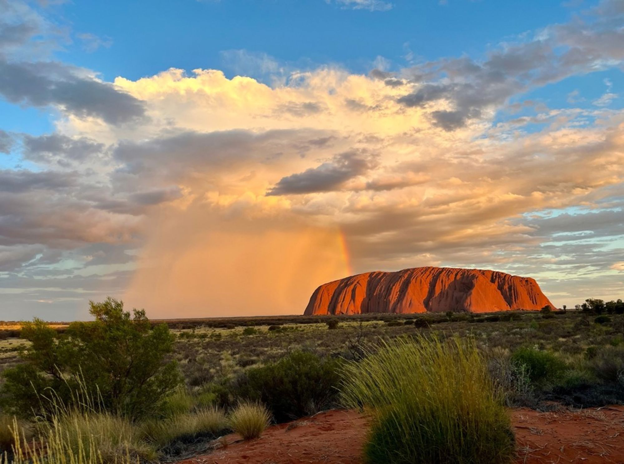 Uluru (Ayers Rock) bij zonsondergang in Australië, met wolkenlucht en regenboog boven het rode monoliet.