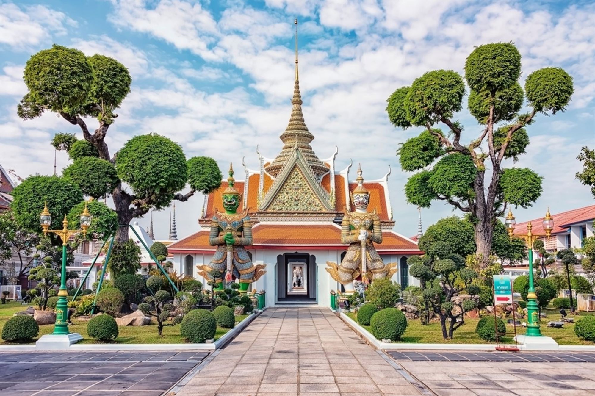Ingang van Wat Arun-tempel in Bangkok met twee wachtersbeelden en gesnoeide bomen.