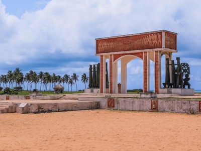 Monumentale poort met reliëfs en beelden aan het strand, met palmbomen en zee op de achtergrond.
