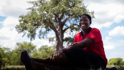 Phyllis uit Zimbabwe zit glimlachend in het gras, met een grote boom op de achtergrond.