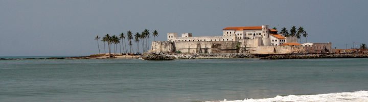 Fort Elmina aan de kust van Ghana, met palmbomen en zee op de voorgrond.